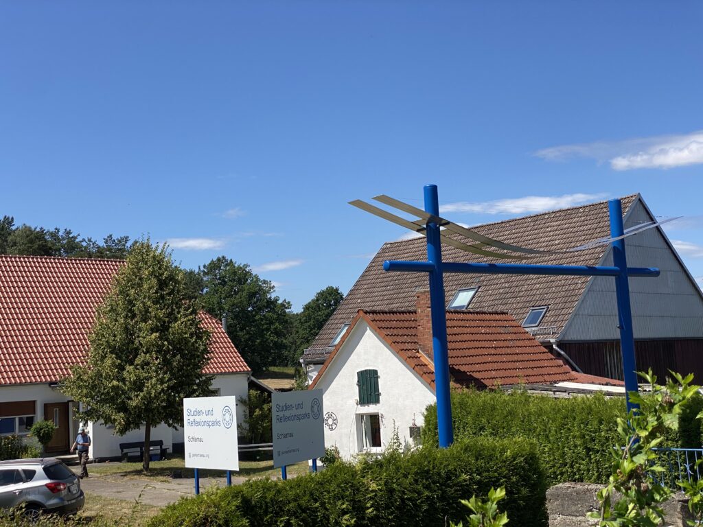 Auf dem Foto sieht man das Eingangstor des Studien- und Reflexionspark Schlamau. Es besteht aus 2 aufrechten Stangen und einer Querstange in der Farbe blau, die sich in ca. 4 Meter Höhe befindet. Über der Querstange ist ein geschwungener Edelstahlbogen sichtbar. Der Edelstahlbogen deutet die Form einer Kugel an. Die Form des Tores erinnert an ähnliche Tore aus dem Shintoismus in Japan.