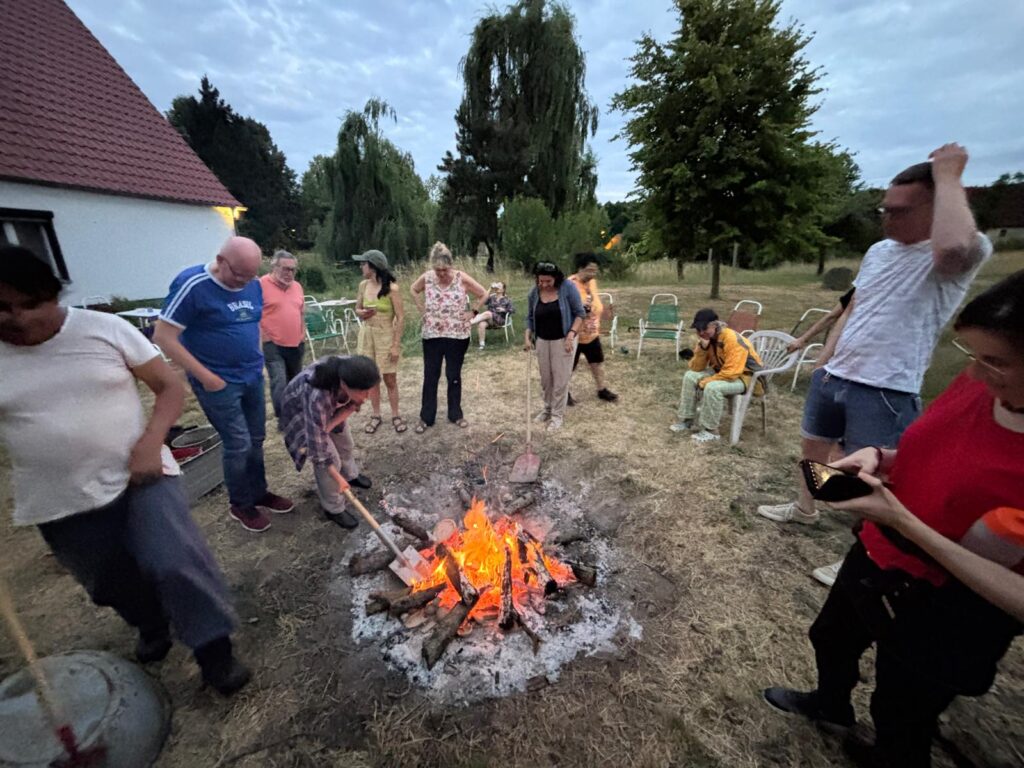 Auf dem Foto sieht man einen Gruppe Menschen zwischen 30 und 50 Jahren alt, beiderlei Geschlechts. Sie stehen um ein Lagerfeuer, eine Person macht etwas mit einem Spaten im Feuer, eine andere Person wartet mit einer Schaufel neben dem Feuer, einen dritte sitzt und schaut ins Feuer. Links im Bild ist die Ecke eines Hauses zu sehen, im Hintergrund Bäume. Das Foto entstand in einem Workshop der „Keramik & Meditation“ hieß und bei dem kleinen Objekt aus Ton mit Feuer gebrannt wurden. Diese Technik heißt Raku.