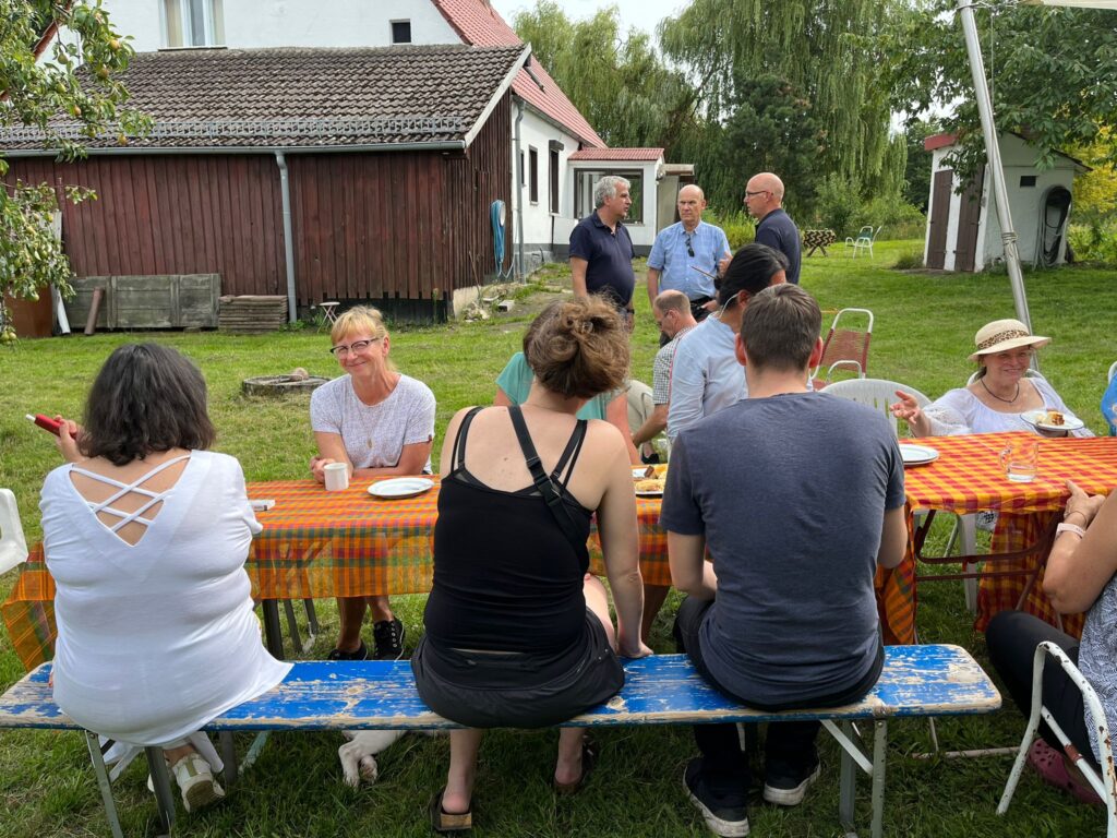 Vordergrund des Fotos sieht man einen aufgestellte Biertisch-Garnitur, die mit einer Orange-grünen Tischdecke gedeckt ist. Ungefähr 10 Menschen sitzen auf den Bänken und erzählen sich gegenseitig etwas. Die Umgebung ist sehr grün, alles sieht sehr sommerlich aus. Im Hintergrund sieht man den Teil eines alten Bauernhauses, welches weiße Wände und ein rotes Dach hat. Davor stehen 3 Männer und unterhalten sich eher ernst.